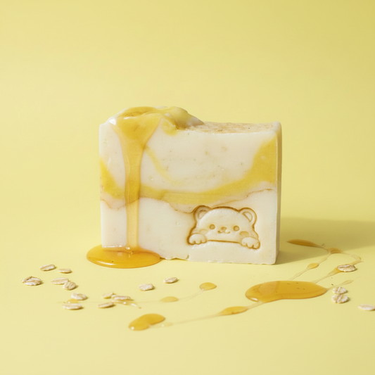 A rectangular bar of handmade soap with a white base, yellow honey swirls, and a carved bear face, set against a yellow background with honey drizzle and oats scattered around.